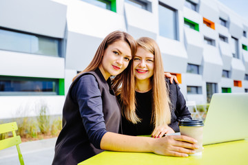 Two female students hugging and looking at camera while sitting in cafe terrace outdoors. Education, lifestyle and business concept.