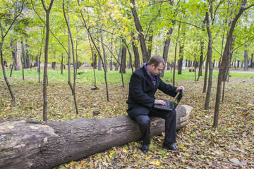Fototapeta premium Businessman having a laptop in the park in the fall. A man is working at a computer in the park.