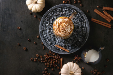 Obraz na płótnie Canvas Glass of spicy pumpkin latte with whipped cream and cinnamon standing on black ornate serving board with decorative white pumpkins . Coffee beans and spices above. Dark background. Top view, space