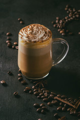 Glass of spicy pumpkin latte with whipped cream and cinnamon standing on black serving board. Coffee beans and spices above. Dark background.
