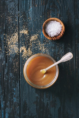 Glass jar of homemade salted caramel sauce with spoon, brown sugar and bowl of salt. Over old dark...