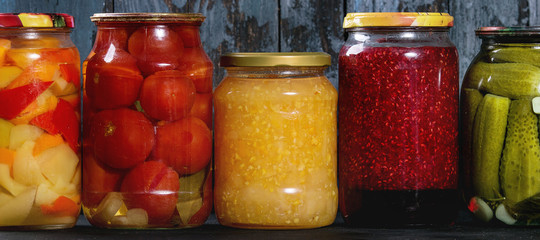 Variety glass jars of homemade pickled or fermented vegetables and jams in row with old dark blue wooden plank background. Seasonal preserves.