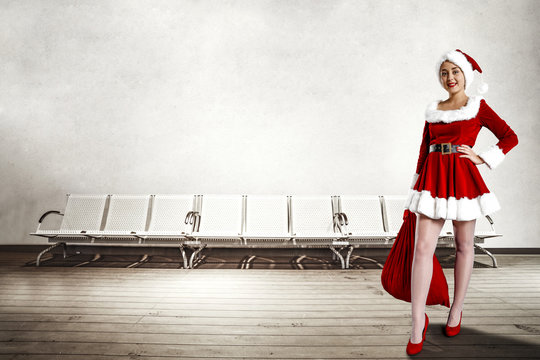 Santa claus woman in red dress and white pantyhose. He holds a bag of holy Santa Claus. It stands on a wooden floor. Background of white metal chairs and white wall.