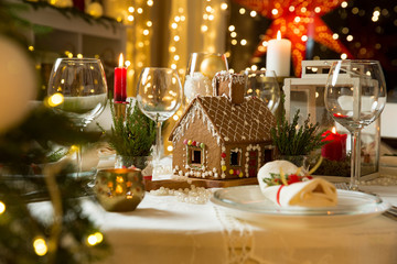 Beautiful served table with decorations, candles and lanterns. Little gingerbread house with glaze on white tablecloth. Living room decorated with lights and Christmas tree. Holiday setting