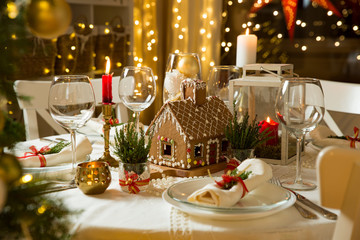 Beautiful served table with decorations, candles and lanterns. Little gingerbread house with glaze on white tablecloth. Living room decorated with lights and Christmas tree. Holiday setting
