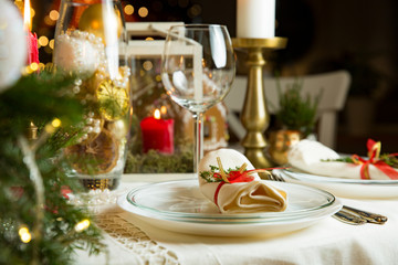 Beautiful served table with decorations, candles and lanterns. Little gingerbread house with glaze on white tablecloth. Living room decorated with lights and Christmas tree. Holiday setting close up