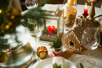 Obraz na płótnie Canvas Beautiful served table with decorations, candles and lanterns. Little gingerbread house with glaze on white tablecloth. Living room decorated with lights and Christmas tree. Holiday setting close up