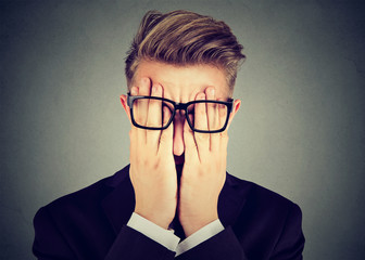 Closeup portrait young man in glasses covering face eyes with both hands