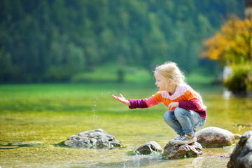 Adorable girl playing by Konigssee lake in Germany on warm summer day. Cute child having fun feeding ducks and throwing stones into the lake. - obrazy, fototapety, plakaty