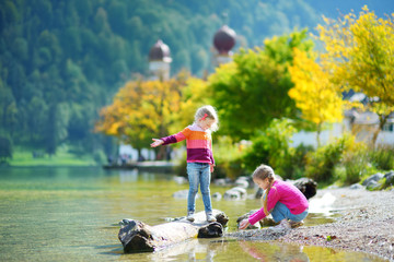 Adorable sisters playing by Hallstatter See lake in Austria on warm summer day. Cute children having fun splashing water and throwing stones into the lake.