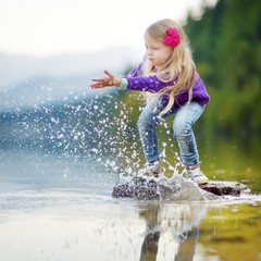 Adorable girl playing by Hallstatter See lake in Austria on warm summer day. Cute child having fun splashing water and throwing stones into the lake.