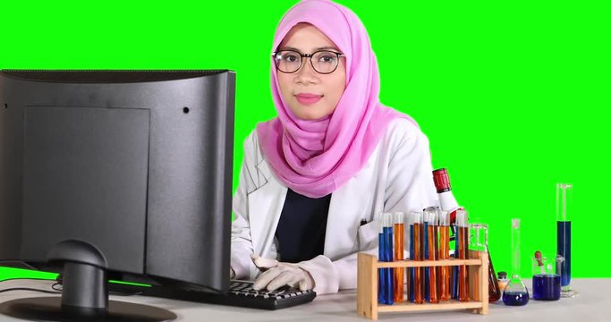 Female scientist using a computer and smiling at the camera with test tube on the table. Shot in 4k resolution with green screen background
