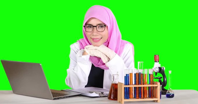 Female scientist with a laptop computer and test tube on the table, smiling at the camera with green screen background. Shot in 4k resolution