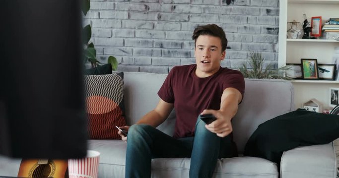Young handsome man turns on the TV set on sofa and uses the phone at home modern design playing remote single time apartment couch laziness looking men couch Shot on RED EPIC camera Slow Motion Shot