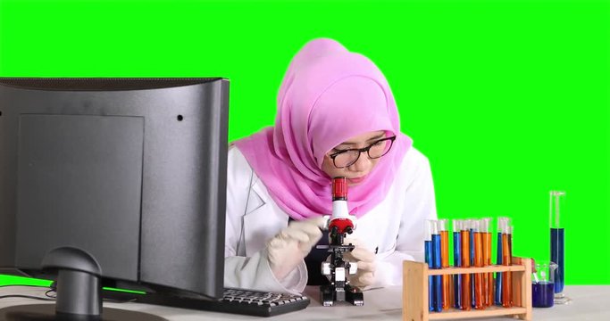 Muslim scientist working with a computer and looking through a microscope with test tube on the table. Shot in 4k resolution with green screen background