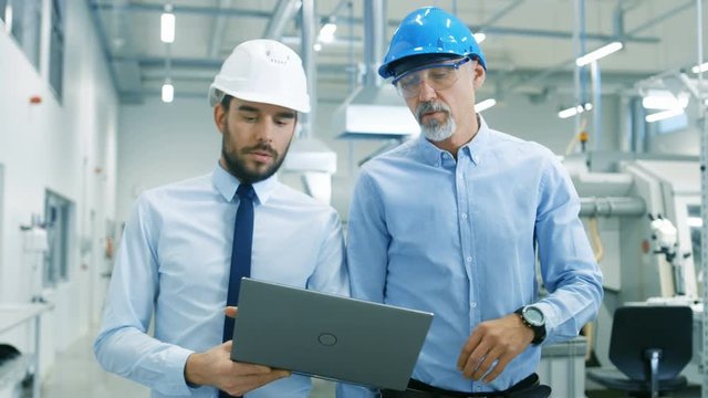 Head of the Project Holds Laptop and  Discusses Product Details with Chief Engineer while They Walk Through Modern Factory. Long Shot.