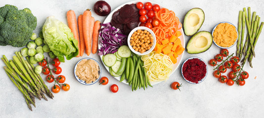 Healthy eating concept, above view of rainbow buddha bowl, various vegetables, carrot, courgette, cabbage, chickpeas, cucumber and tomatoes, on wooden board on white table, long photo, selective focus