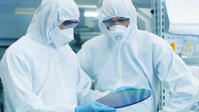 Two Scientists/ Technicians in Sterile Suits Check Semiconductor Silicon Wafer that Reflects Many Different Colors, it will be Made into Computer Chips. 