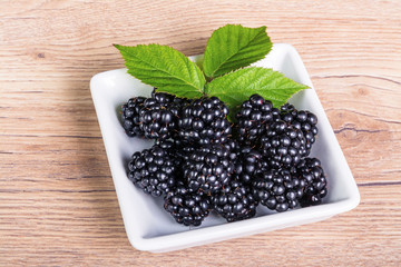 Sweet juicy blackberries in white squared bowl on wooden background. Tasty fresh fruit with decorative green leaves on brown garden table.