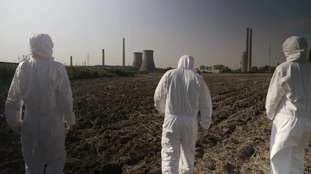 Employees ecologists team in hazmat suits looking at contaminated environment and agricultural area walking towards a nuclear power refinery