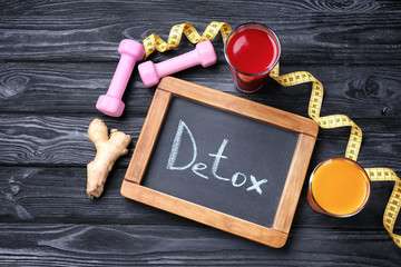 Small chalkboard with word DETOX, fresh juices in glasses and dumbbells on table