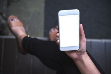 Mockup image of a woman sitting crossed leg and holding white mobile phone with blank screen with concrete floor background