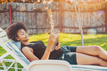 Attractive young Brazilian lady laying on lounge and chatting with friend via tablet pc; calm black...