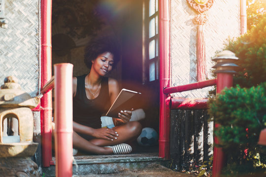 Cute young black curly student girl is sitting on the floor inside of pagoda with red beams, with legs crossed and thoughtfully reading digital book on her tablet pc, warm summer sunny day