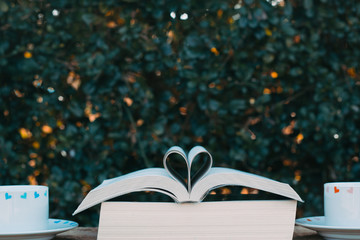 An open book in heart shape and two cups of coffee on a wooden chair in the park with green leaves...