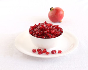Pomegranate seeds in a white ceramic bowl on a plate with a few spilled seeds.