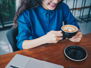 Fototapeta na wymiar Young asian woman sitting in coffee shop at wooden table, hand holding coffee cup, Using laptop browsing internet, chatting, blogging, shopping onlie.