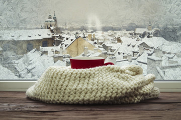 Fototapeta na wymiar Frozen window and red cup with hot coffee and scarf on the podkonneke with a beautiful view of winter Prague