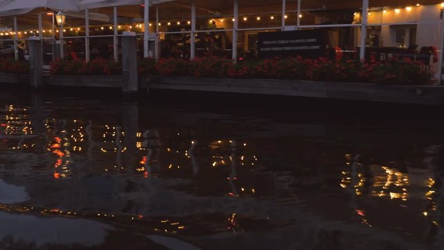 Lights reflects beautifully off the gently moving waters of a canal in Amsterdam. The tables are still mostly empty during the early evening hours.