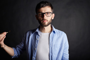 Close up portrait of attractive young serious or dangerous Caucasian man with burning match on his hand on black studio wall. Brunette hipster in trendy spectacles posing indoors with fire in his hand