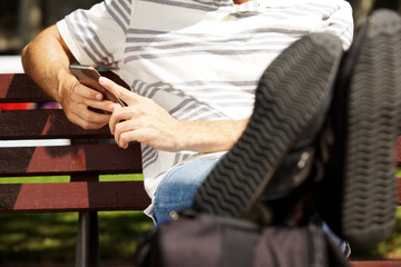 Close up man sitting on bench outdoors with legs on bag and using mobile phone