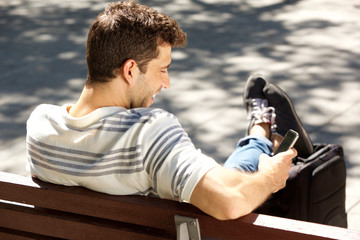 Smiling young man sitting on bench outdoors with bag and using smart phone