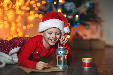 Boy writing letter to Santa Claus in red hat near the Christmas tree