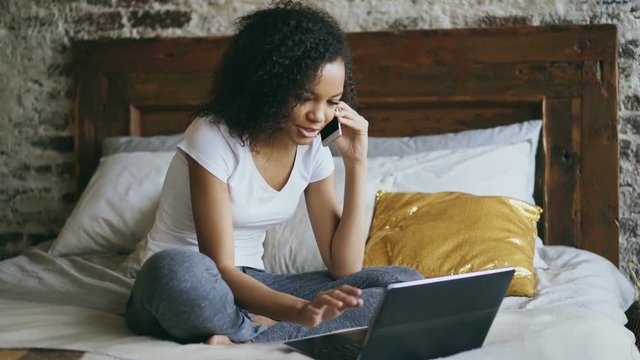 Curly african american girl talking smartphone and using laptop computer sitting in bed at home