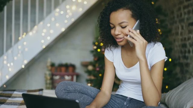 Curly mixed race girl chatting smartphone and using laptop computer on Christmas tree backround at home