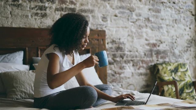 Curly attractive african girl typing on laptop computer for sharing social media sitting in bed at home