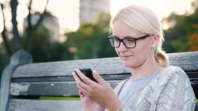 A young woman in glasses is using a smartphone. Sits on a park bench