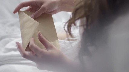 Top view of unrecognizable woman with long hair sitting in bed on early morning and opening envelope with letter from lover