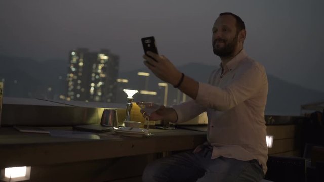Young man with drink taking selfie with cellphone on terrace in bar at night
