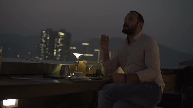 Sad, unhappy man drinking cocktail sitting on terrace in bar at night
