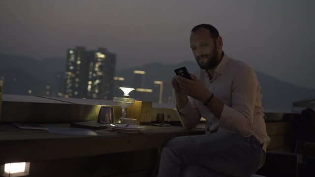 Young man with smartphone drinking cocktail sitting on terrace in bar at night
