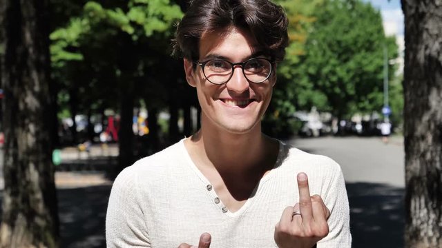 Handsome friendly smiling young man doing screw you sign with middle fingers in a joking way, outdoor in city park