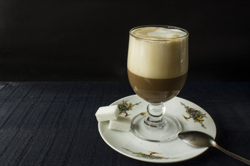 a beautifull glass with coffee latte with high foam of cream and saucer on the black background