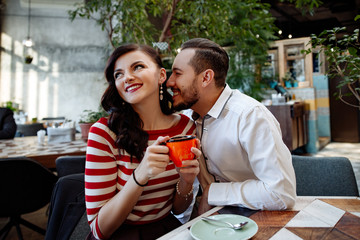date in cafe. The man and the woman communicate smile and laugh behind a cup of coffee. The man whispers to the girl on an ear complements.