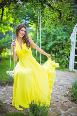 Beautiful girl in yellow dress, riding on swing on background of  blossoming park in the summer.