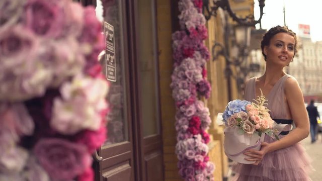attractive young woman in evening dress standing at the entrance to a flower shop. the girl is holding a cardboard hat box with flowers in cancases. a large bouquet of flowers. the girl demanstrates a